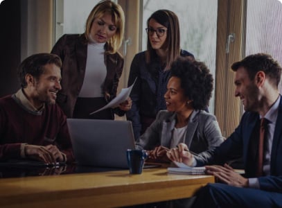 A group of five young professionals are sitting around a table, collaborating on a project. They are looking at a laptop and some documents. The atmosphere is one of focused teamwork.