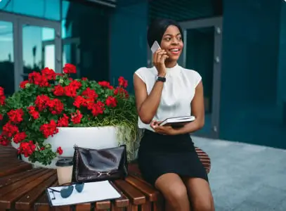 women sitting next to flowes on a bench on the phone, holding a note book