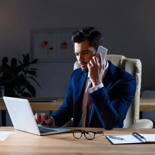 man looking at his computer while talking on the phone, glasses on the side of the desk