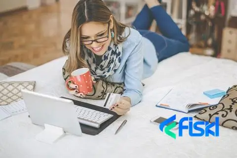 a women holding a mug while lying on a bed looking at her laptop and has a card in her hand wiht the Fiskl logo