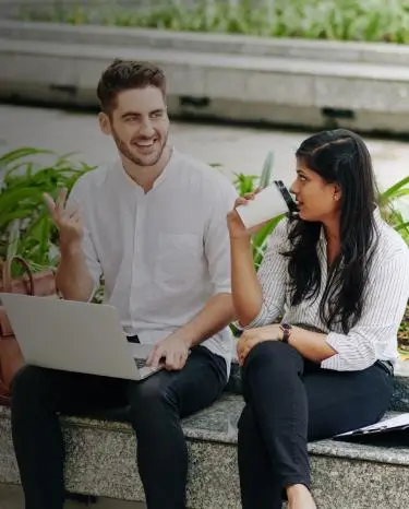 a women and man sitting on a bench outside drinking from cups and working from a laptop