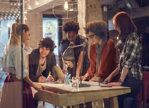 A diverse group of five young professionals collaborating around a desk with a laptop and blueprints in a modern, warmly lit creative office studio.