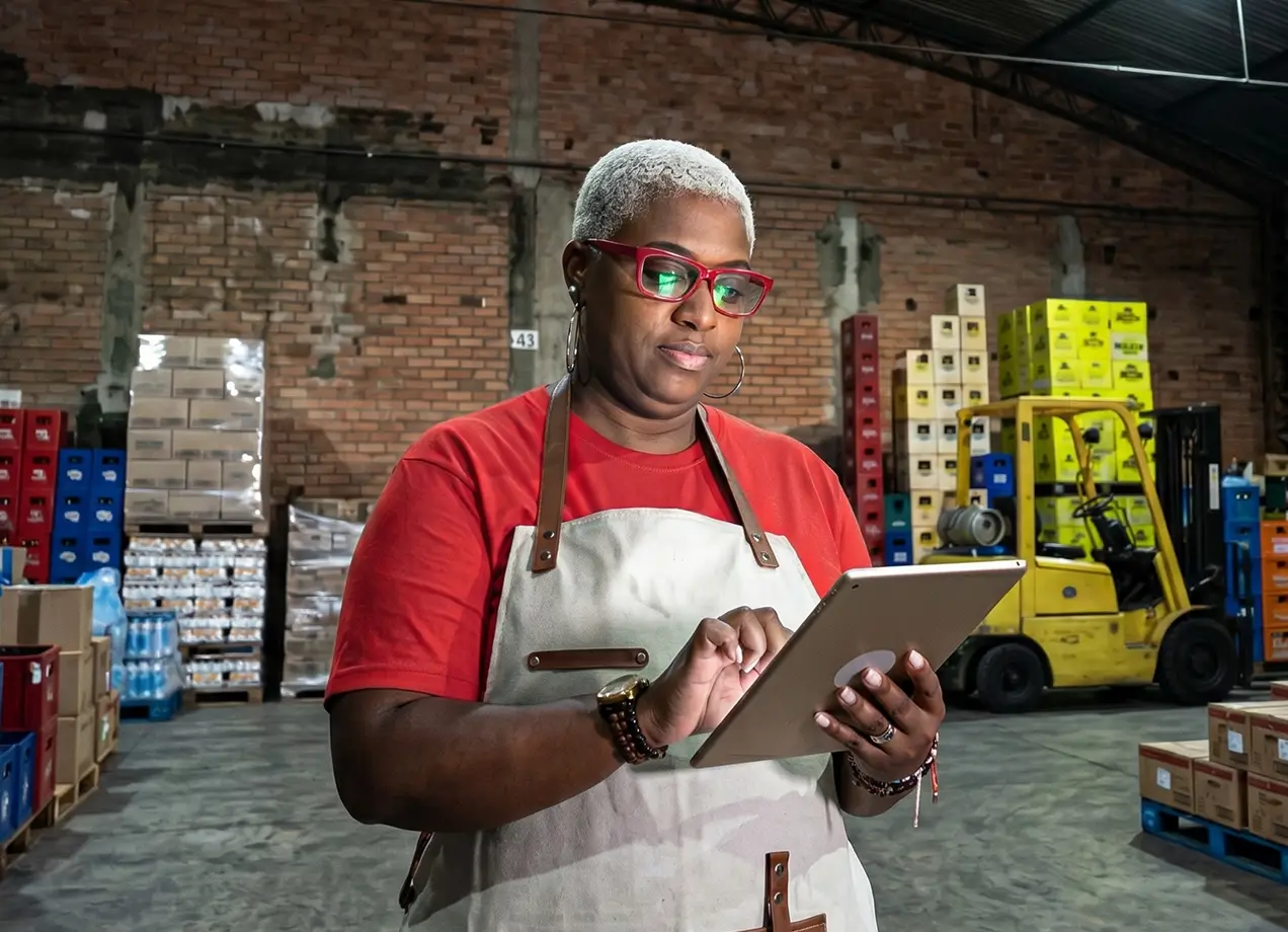 A female warehouse manager with short grey hair and red glasses using a digital tablet in a spacious storage facility filled with pallets of boxed inventory