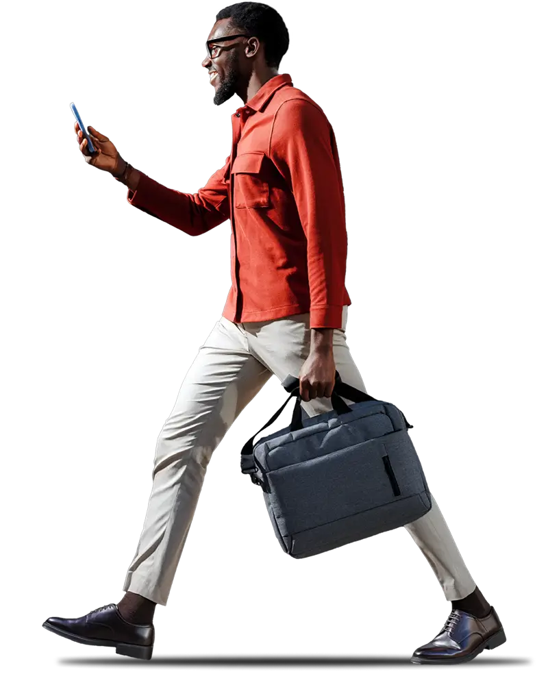 A smiling man in a red button-up shirt and beige trousers walking while looking at his smartphone and carrying a grey laptop briefcase against a black background.