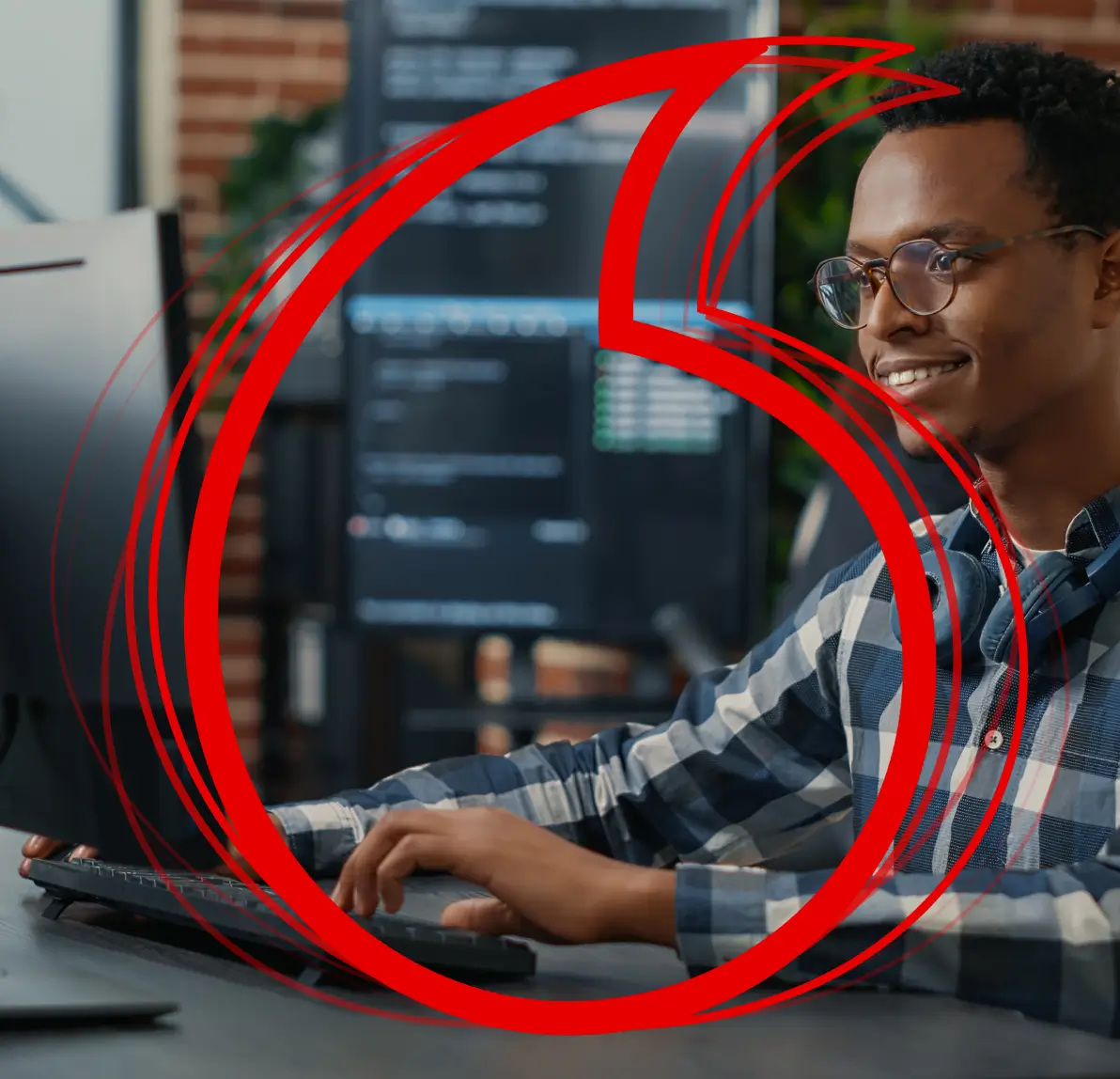 A smiling man wearing glasses works at a computer desk with code on the monitors, overlaid with a large red Vodafone logo.