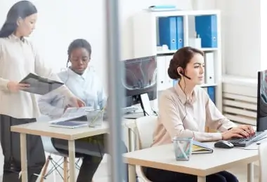 Business professional wearing a headset working on a computer at her desk while colleagues collaborate in the background
