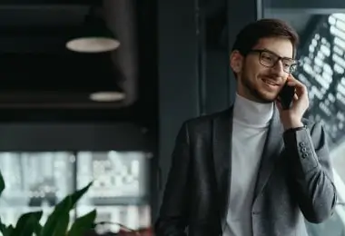 Smiling businessman in a suit and turtleneck talking on his mobile phone near an office window