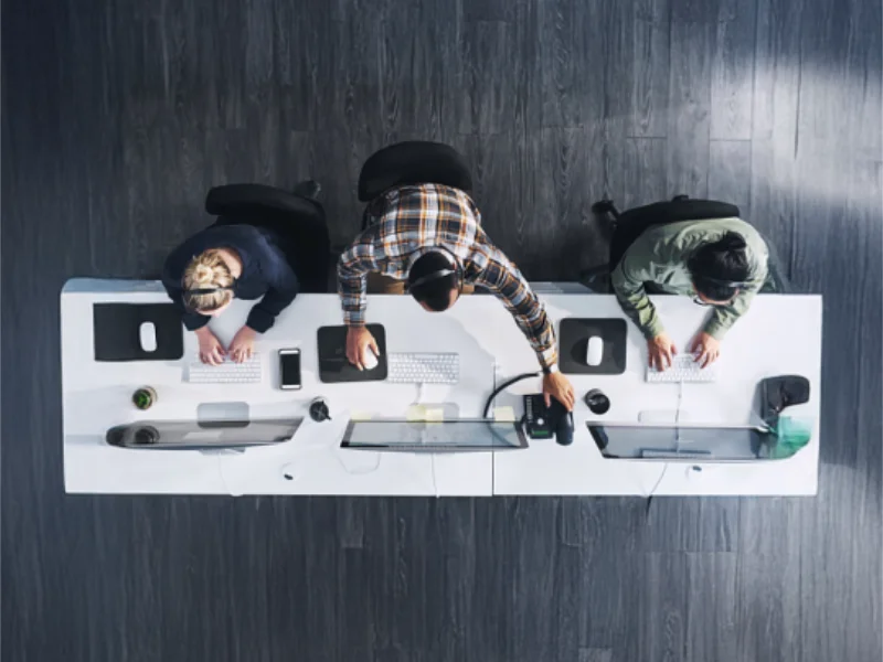 An overhead, high-angle shot of three people working at a white desk in a modern contact centre, typing on keyboards and using computers with headsets on.